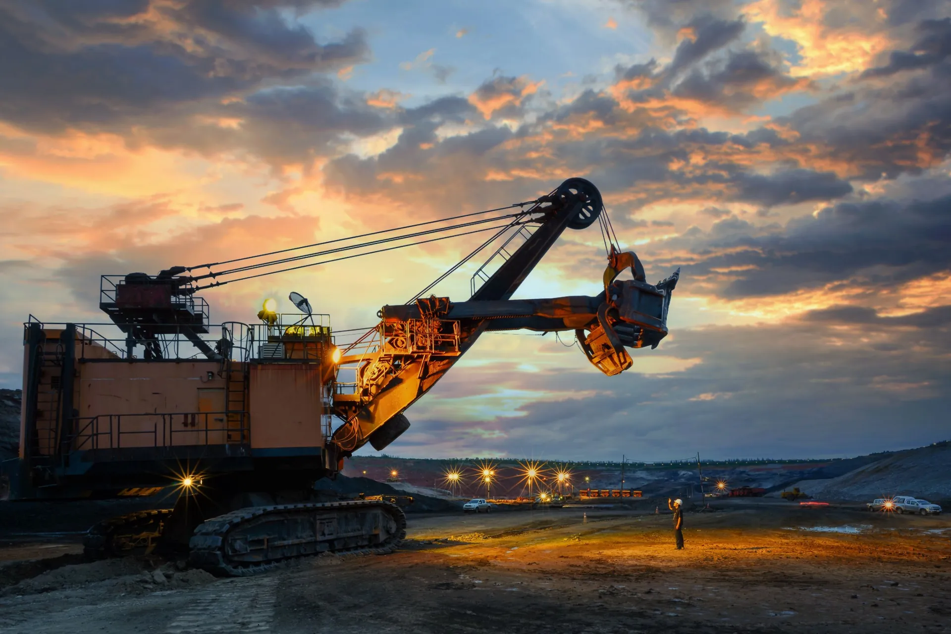 Worker in a hard hat standing beside an electric rope shovel at an open-cut mine at sunset